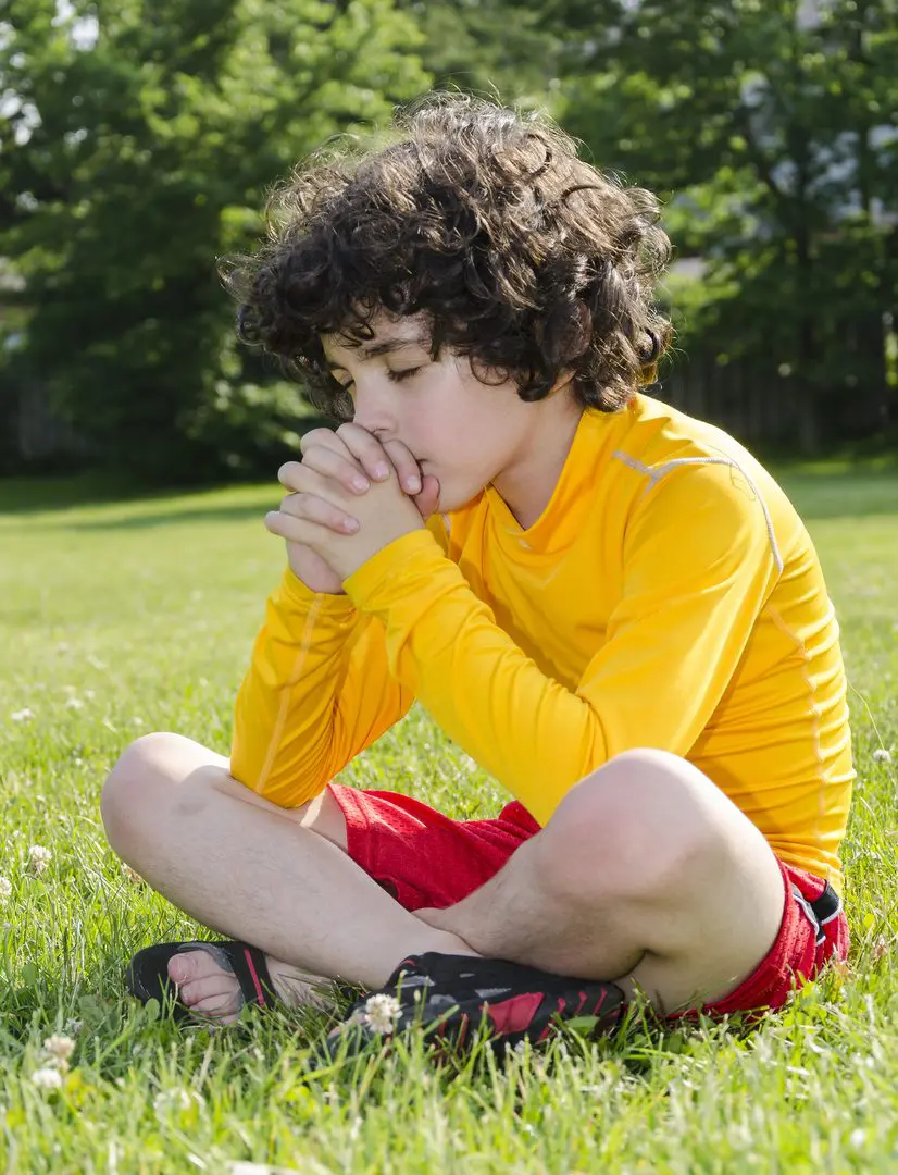 Boy in yellow jacket sitting cross-legged on grass, looking thoughtful.